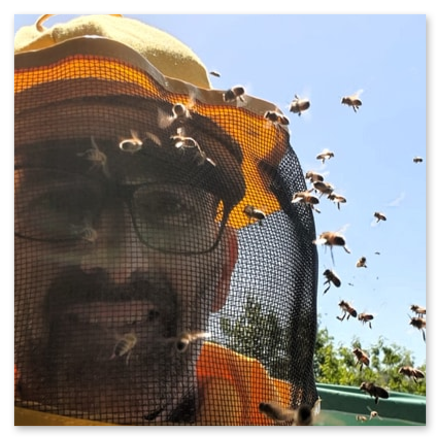 Selfie di Simone Mabellini, Business Coach, con tuta protettiva da apicoltore, rete sul viso e cappello giallo, che sorride mentre uno sciame di api vola contro il cielo azzurro e la vegetazione.